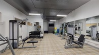 Located at 250 W 5th St in Cincinnati, Ohio, this fitness center offers a comprehensive and well-organized workout facility on a single floor. The space features a neutral color palette of white and dark gray walls, complemented by durable plaid-patterned flooring that adds a subtle design element throughout.

The gym is equipped with an extensive selection of free weights, including a full rack of dumbbells in graduated sizes, accompanied by flat weight benches for a variety of exercises. Multiple selectorized strength machines — including a cable lat pulldown station, a seated leg press, and a Life Fitness multi-function unit — are thoughtfully positioned to allow ample circulation between equipment. For functional training, the space includes kettlebells, stability balls, a BOSU balance trainer, and a medicine ball rack.

Large wall-mounted mirrors run along multiple walls, enhancing the sense of openness and allowing users to monitor their form. Industrial ceiling-mounted fans provide airflow throughout the space, while fluorescent strip lighting ensures bright, even illumination. A wall-mounted flat-screen TV serves as an entertainment focal point on the darker accent wall. The facility is accessed through a dedicated entry door and maintains a clean, purposeful layout suited for a wide range of fitness routines.
