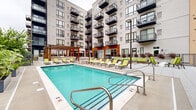 Located at 5245 Wayzata Blvd in St. Louis Park, Minnesota, this outdoor amenity deck offers a well-appointed, resort-inspired environment designed for relaxation and social activity. At the heart of the space is a generously sized swimming pool with crystal-clear turquoise water, framed by a wide pool deck lined with bright chartreuse lounge chairs and accent seating. Adjacent to the main pool, a separate spa pool provides a more intimate retreat, accompanied by a private pergola-covered cabana nook outfitted with cushioned chaise lounges. A series of slatted wood cabanas with striped cushioned daybeds offer shaded, semi-private lounging areas, blending natural wood tones with a modern architectural aesthetic. The outdoor social area features an open-air dining space with tables, chairs, and large cantilever umbrellas, set beneath a pergola structure with accent lighting. A bocce ball court adds a recreational element to the courtyard. Lush greenery, raised planters with colorful flowers, and mature trees soften the hardscape throughout. The deck is enclosed by a wrought iron fence, with the surrounding multi-story residential building providing a sense of enclosure and community. The overall design balances contemporary style with approachable, functional outdoor living.
