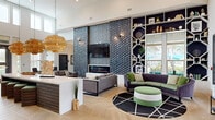 Located at Crest Preserve in Bonita Springs, Florida, this single-floor community clubhouse offers a generously sized, thoughtfully designed gathering space. The centerpiece of the interior is a dramatic open-concept lounge featuring soaring ceilings, light-toned chevron-patterned hardwood flooring, and a striking textured accent wall finished in deep charcoal tile. A sleek linear fireplace is flanked by a wall-mounted television, while an eye-catching built-in shelving unit with geometric cutouts adds architectural interest. Woven rattan pendant lights hang above a central island with bar-height seating, casting warm ambient light throughout the space. Multiple seating arrangements — including modular sofas and a curved sectional with a round ottoman — invite social interaction and relaxation. Natural light pours in through large windows and glass double doors that open toward an outdoor pool area. A cozy reading nook features tall windows, plush armchairs, and a compact library station. A dedicated dining area with a glass-paned office window provides a semi-private workspace setting. The facility also includes a secure package receiving room equipped with wall-mounted wire shelving for organized parcel storage. The overall ambiance is modern and welcoming, blending contemporary design with warm, resort-inspired details.