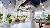 Located at 100 Commons Park N in Stamford, Connecticut, this single-floor lobby space delivers a striking industrial-chic aesthetic that blends modern design with urban character. Soaring ceilings with exposed silver ductwork and bold blue-painted soffits set a dramatic tone, while pendant lighting with cage and dome fixtures casts a warm, inviting glow throughout the open floor plan.

Floor-to-ceiling glass curtain walls wrap the perimeter, flooding the interior with abundant natural light and offering panoramic views of the surrounding Stamford cityscape. Polished concrete floors anchor the space, complementing the raw, industrial elements above.

A welcoming lounge area features quilted yellow accent chairs, a patterned area rug, and lush tropical greenery, creating an energetic yet comfortable gathering space. Colorful artwork and bold "NV" signage add a curated, urban personality to the walls.

A separate reception area showcases a standout steampunk-inspired design, with a riveted metallic front desk, corrugated metal walling, and an eye-catching arrangement of gear-framed circular mirrors in varied sizes and finishes. Industrial pendant lights complete this unique focal point.

High-top communal tables near the glazed façade offer additional seating options, making this lobby a versatile and design-forward space suited for both welcoming visitors and informal gatherings.