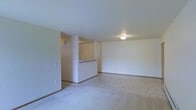 This single-floor residence offers a clean and functional layout designed for comfortable everyday living. The main living area is spacious and open, finished with soft, neutral-toned carpeting that extends across the floor and contributes to a calm, understated ambiance. The walls are painted in a crisp white, creating a bright and airy atmosphere throughout the space.

A defining architectural feature is the half-wall pass-through counter that separates the living area from the kitchen, allowing for an open feel while maintaining a sense of organization between the two spaces. The kitchen area, visible beyond the pass-through, features hard flooring that contrasts with the carpeted living area, providing a practical transition between spaces.

Flush-mounted ceiling lighting provides consistent, even illumination throughout the room, keeping the interior well-lit and inviting. Structural columns near the pass-through add an interesting architectural element to the otherwise streamlined interior. The overall design is simple and versatile, offering a neutral canvas that is well-suited for a variety of furnishing styles and personal touches. Clean lines, minimal detailing, and a cohesive color palette make this a practical and welcoming single-floor living space.