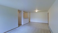 This single-floor residence offers a clean and functional layout designed for comfortable everyday living. The main living area is spacious and open, finished with soft, neutral-toned carpeting that extends across the floor and contributes to a calm, understated ambiance. The walls are painted in a crisp white, creating a bright and airy atmosphere throughout the space.

A defining architectural feature is the half-wall pass-through counter that separates the living area from the kitchen, allowing for an open feel while maintaining a sense of organization between the two spaces. The kitchen area, visible beyond the pass-through, features hard flooring that contrasts with the carpeted living area, providing a practical transition between spaces.

Flush-mounted ceiling lighting provides consistent, even illumination throughout the room, keeping the interior well-lit and inviting. Structural columns near the pass-through add an interesting architectural element to the otherwise streamlined interior. The overall design is simple and versatile, offering a neutral canvas that is well-suited for a variety of furnishing styles and personal touches. Clean lines, minimal detailing, and a cohesive color palette make this a practical and welcoming single-floor living space.