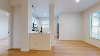 This single-floor residence presents a clean, modern aesthetic defined by light wood flooring and crisp white walls throughout. The open-concept layout connects the kitchen and living area seamlessly, creating a bright, cohesive space. The kitchen is well-appointed with white shaker-style cabinetry accented by matte black hardware, a herringbone marble-look tile backsplash, white quartz countertops, and a full suite of stainless steel GE appliances including a range, over-the-range microwave, dishwasher, and refrigerator. A peninsula with a sink adds additional prep space and casual dining potential.

The living area benefits from large windows fitted with horizontal blinds, offering views of lush greenery. The bedrooms continue the light, airy feel with the same wood flooring and windows framing tree canopy views. A spacious walk-in closet with wire shelving provides generous storage. The bathroom features a tub-shower combination enclosed by a glass panel, large-format marble-look tile surround, white vanity cabinetry with black hardware, and warm lighting. A dedicated laundry room houses a full-size front-loading washer and dryer with wire shelving above.