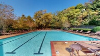 This outdoor aquatic facility centers around a generously sized lap pool with clearly marked swim lanes and a classic brick coping surround. The crystal-clear turquoise water is complemented by a wide concrete deck lined with rows of sling-style lounge chairs, offering ample space for sunbathing and relaxation. A covered pavilion runs alongside the pool area, supported by crisp white columns and featuring a paneled ceiling that provides welcome shade. The pavilion is furnished with wrought-iron bistro tables and additional lounge seating, making it an ideal spot to unwind out of direct sun. The structure's exterior is finished in a soft blue-gray horizontal shiplap siding with white louvered vents, lending a clean, classic aesthetic. Framed informational postings and pool rules are displayed along the pavilion wall, reflecting the facility's well-organized management. The entire space is enveloped by a dense canopy of mature deciduous trees, which provide a natural, park-like setting and a sense of privacy. The combination of open sky, abundant greenery, and thoughtfully arranged amenities creates a serene and inviting atmosphere suited for both lap swimming and leisurely outdoor recreation.