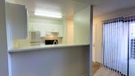 This single-floor residence offers a practical and efficiently designed layout that makes thoughtful use of its available space. The kitchen serves as a central feature, showcasing clean white shaker-style cabinetry that extends to the upper walls, providing ample storage. Stainless steel appliances, including a refrigerator and a range with an overhead ventilation hood, add a modern touch to the cooking area. A peninsula counter with a light-toned surface extends outward, creating a natural dividing point between the kitchen and the adjacent living or entry area, while also offering additional prep and dining space. The kitchen benefits from bright overhead fluorescent lighting that illuminates the workspace effectively. Textured ceilings are present throughout, adding a subtle tactile quality to the interior. The entry area, visible from the kitchen, features wood-look flooring and provides access to additional rooms. A smaller secondary space, likely a closet or utility room, is fitted with a ceiling-mounted light fixture and built-in shelving, maximizing storage efficiency. The overall interior palette is light and neutral, with soft greens and whites creating a calm, welcoming ambiance throughout the home.