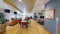 Located at 2775 S Brook Dr in Denver, Colorado, this thoughtfully designed single-floor community facility offers a warm and functional environment across several distinct yet interconnected common areas. The entry foyer makes a striking first impression with warm hardwood-style flooring, a decorative glass water feature partition, and oversized potted greenery that frames the transition into the main living spaces. Large glass doors and floor-to-ceiling windows flood the interior with natural light and provide views of the surrounding outdoor landscape. The main lounge area features generous seating arrangements, bold drum pendant lighting, a television, and an upright piano, creating a versatile gathering space suited for both relaxation and entertainment. Adjacent to the lounge, a cozy library and media alcove showcases built-in shelving stocked with books, comfortable upholstered seating, and a stone-clad fireplace wall with a mounted flat-screen television beneath a coffered ceiling detail. The dining and social area offers communal tables and chairs, bathed in natural light from sheer-curtained windows with direct access to an outdoor patio. A fully equipped community kitchen anchors the space with a large central island with bar seating, mosaic tile backsplash, pendant lighting, full appliances, and ample cabinetry—ideal for communal cooking and events. Throughout, the consistent use of warm wood flooring, neutral tones, and modern furnishings creates a cohesive, inviting ambiance.