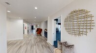 Located at 1000 Navaho Dr in Raleigh, NC, this single-floor amenity space offers a well-designed layout that combines functionality with a refined aesthetic. The hallways serve as more than simple passageways — finished with light wood-look flooring, recessed lighting, and tasteful decorative accents including a striking gold lattice wall piece, they create a welcoming corridor that connects the various areas of the space. Comfortable seating, including an upholstered accent chair and a cushioned bench with decorative pillows, adds a lounge-like quality to the hallways. A built-in navy cabinetry unit provides additional character and storage along the corridor. A dedicated dining or meeting area is also visible, featuring a long white table surrounded by neutral-toned chairs beneath bright recessed lighting. The standout feature of this space is the expansive exercise room, which boasts soaring vaulted ceilings with dramatic black upper walls contrasting against crisp white trim. Large windows flood the room with natural light and offer outdoor views. The fitness area is fully equipped with treadmills, stationary bikes, cable machines, a squat rack, weight benches, a full dumbbell rack, stability balls, and wall-mounted mirrors — providing a comprehensive workout environment.