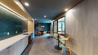 This contemporary amenity space is thoughtfully designed to serve a variety of social and professional needs. The main lounge area features large-format tile flooring, dark teal-painted ceilings, and warm light wood wall paneling, creating a refined yet inviting atmosphere. A built-in kitchenette with a sink and ample counter space anchors one side, complemented by floor-to-ceiling glass partitions that draw in natural light and provide views to adjacent outdoor areas. Flexible seating options abound, including high-top tables with bold yellow bar stools and a round black dining table with modern wood chairs beneath a sculptural pendant light fixture.

Transitioning deeper into the space, the design shifts dramatically with striking, black-and-white patterned wallcoverings that lend a bold artistic character to the interior rooms. A dedicated media lounge features a large wall-mounted flat-screen television and a generously sized sectional sofa on green carpet flooring — ideal for relaxed viewing or social gatherings. Adjacent to this is a focused work area equipped with built-in light wood desks, individual workstations with privacy dividers, floating shelving, and black task chairs, offering a productive environment within the same footprint.