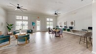 This well-appointed office suite occupies a single floor and is thoughtfully designed to balance professional functionality with an inviting, contemporary aesthetic. Light hardwood flooring runs throughout the space, complementing the bright white walls and crown molding that lend a polished, cohesive feel.

The main open-plan office area is generously proportioned, featuring high ceilings with ceiling fans, large windows with transom lights above, and glass-paned French doors that flood the space with natural light and offer views of lush surrounding greenery. This area includes a central conference table, a dedicated computer workstation counter along one wall, and a comfortable lounge seating arrangement with teal accent chairs, creating a versatile environment for both collaboration and focused work.

The second office is a more intimate, private space distinguished by soaring ceilings with clerestory windows and a warm wood desk paired with a lounge seating area featuring plush gray chairs. Curated abstract artwork adorns the walls throughout both offices, adding a refined artistic touch.

A welcoming reception or lounge area connects the two offices, outfitted with cognac leather accent chairs and soft neutral tones. The space benefits from ample natural light, a wall-mounted mini-split system, and thoughtful design details that create a calm, professional ambiance ideal for a modern office environment.