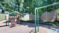 This outdoor recreational space features a well-equipped playground area designed for active play. A multi-component play structure with red metal framing includes a slide, climbing elements, and a tic-tac-toe panel, set atop a safety surface of wood chip mulch that cushions the play zone. Adjacent to the structure stands a green metal swing set frame, providing additional play options. Mature deciduous and evergreen trees border the space, offering natural shade across the grounds during warmer months. A stretch of open, manicured green lawn extends beyond the playground, providing ample room for recreational activities. The area is enclosed by a tall wooden privacy fence, creating a defined and secure boundary. Residential buildings are visible in the background, indicating the playground serves as a shared amenity within a surrounding residential community. The overall setting is calm and well-maintained, combining structured play equipment with open natural space in a comfortable, shaded outdoor environment.