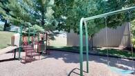 This outdoor recreational space features a well-equipped playground area designed for active play. A multi-component play structure with red metal framing includes a slide, climbing elements, and a tic-tac-toe panel, set atop a safety surface of wood chip mulch that cushions the play zone. Adjacent to the structure stands a green metal swing set frame, providing additional play options. Mature deciduous and evergreen trees border the space, offering natural shade across the grounds during warmer months. A stretch of open, manicured green lawn extends beyond the playground, providing ample room for recreational activities. The area is enclosed by a tall wooden privacy fence, creating a defined and secure boundary. Residential buildings are visible in the background, indicating the playground serves as a shared amenity within a surrounding residential community. The overall setting is calm and well-maintained, combining structured play equipment with open natural space in a comfortable, shaded outdoor environment.