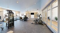 Located at 95 Newport Ave in East Providence, Rhode Island, this single-floor amenity space offers a well-appointed fitness and common area designed with a clean, contemporary aesthetic. The layout spans a generous footprint, seamlessly connecting bright, open corridors to a dedicated exercise room.

The corridors feature warm light wood flooring, soaring ceilings, and an abundance of natural light streaming through large windows. Seating areas with modern accent chairs and decorative greenery are thoughtfully placed along the hallway, creating welcoming transitional spaces. A mezzanine-style railing detail adds architectural interest and visual depth.

The exercise room is the centerpiece of the space, outfitted with a comprehensive selection of fitness equipment including treadmills, elliptical machines, cable and weight machines, a free weight rack with dumbbells, and a bench press station. Rubber flooring provides durability and safety underfoot, while large floor-to-ceiling mirrors and expansive windows enhance the sense of openness and bring in natural light. Vaulted ceilings give the room a spacious, airy feel, and a wall-mounted flat-screen television adds a modern convenience. The overall ambiance is bright, clean, and professional, making it a fully functional fitness facility suited for a variety of workout routines.