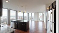 This contemporary single-floor residence offers a thoughtfully designed open-concept layout highlighted by striking floor-to-ceiling windows that frame panoramic waterfront and city views. Rich hardwood flooring runs throughout the main living and kitchen areas, creating a warm contrast against the clean white walls and modern finishes.

The kitchen serves as a central hub of the space, anchored by a large island with granite countertops and pendant lighting overhead. Dark espresso cabinetry pairs with a full suite of stainless steel appliances, including a refrigerator, range, microwave, and dishwasher. The open layout allows the kitchen to flow seamlessly into the generous living area, which is bathed in natural light from the expansive window wall.

The bedrooms are carpeted for comfort and include standard closets with built-in shelving. Each room receives natural light through well-positioned windows with urban views. Two bathrooms serve the unit: one features a walk-in glass-enclosed shower with dark vanity cabinetry, while the second includes a full soaking tub and shower combination. The overall aesthetic is clean, modern, and functional throughout.