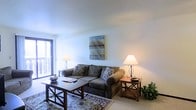 This single-floor space presents a comfortable and inviting living area designed with a clean, understated aesthetic. The living room is anchored by a plush, dark gray sofa accented with decorative pillows, paired with a classic wooden coffee table and coordinating side tables, each topped with elegant table lamps that cast a warm, ambient glow. A patterned area rug adds texture and visual interest to the floor, complementing the neutral tones of the room. Wall décor includes a landscape-inspired canvas painting, contributing a subtle artistic touch to the otherwise minimalist walls. A large sliding glass door or window along one wall allows natural light to filter into the space, brightening the interior and providing a connection to the outdoors. The smooth, light-colored walls and ceiling create an open, airy feel throughout. The overall ambiance is relaxed and residential, with thoughtful furnishing choices that balance comfort and style, making the living area a welcoming and functional space for everyday use.