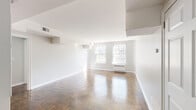 This single-floor space showcases a thoughtfully designed layout with a bright, airy atmosphere defined by crisp white walls and polished parquet wood flooring throughout the living areas. The herringbone-patterned parquet floors add a timeless, classic character to the space, catching natural light and lending warmth to each room.

The expansive main living area benefits from multiple double-hung windows that allow generous natural light to fill the space. Architectural details include stepped ceiling soffits and built-in HVAC units beneath the windows, providing efficient climate control. A secondary room, finished in soft gray tones, also features parquet flooring and a large window with views of surrounding greenery and neighboring buildings.

The kitchen is notably updated, featuring veined granite countertops in cool gray and white tones, white shaker-style cabinetry, stainless steel appliances including a dishwasher, and a stainless undermount sink. A large window overlooks lush, blooming trees, making this workspace especially pleasant. The kitchen is finished with marble-look tile flooring.

The bathroom is clean and functional, featuring classic white subway tile wainscoting, a soaking tub, pedestal-style vanity, mosaic tile flooring, and a window for natural ventilation and light.