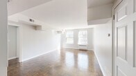 This single-floor space showcases a thoughtfully designed layout with a bright, airy atmosphere defined by crisp white walls and polished parquet wood flooring throughout the living areas. The herringbone-patterned parquet floors add a timeless, classic character to the space, catching natural light and lending warmth to each room.

The expansive main living area benefits from multiple double-hung windows that allow generous natural light to fill the space. Architectural details include stepped ceiling soffits and built-in HVAC units beneath the windows, providing efficient climate control. A secondary room, finished in soft gray tones, also features parquet flooring and a large window with views of surrounding greenery and neighboring buildings.

The kitchen is notably updated, featuring veined granite countertops in cool gray and white tones, white shaker-style cabinetry, stainless steel appliances including a dishwasher, and a stainless undermount sink. A large window overlooks lush, blooming trees, making this workspace especially pleasant. The kitchen is finished with marble-look tile flooring.

The bathroom is clean and functional, featuring classic white subway tile wainscoting, a soaking tub, pedestal-style vanity, mosaic tile flooring, and a window for natural ventilation and light.