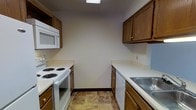 This single-floor space offers a practical and well-organized layout designed for comfortable everyday living. The kitchen is laid out in a classic galley configuration, with cabinetry and countertops lining both sides to maximize efficiency and workflow. Warm-toned wood cabinetry provides generous storage both above and below the counters, lending the kitchen a cozy and traditional feel. White appliances, including a coil-burner electric range, an over-the-range microwave, a dishwasher, and a double-basin stainless steel sink, are neatly integrated into the layout. The light-colored countertops provide ample prep space, complementing the neutral wall tones that keep the space feeling open and clean. Durable tile flooring in earthy, natural tones runs through the kitchen, adding texture and practicality underfoot. Overhead lighting ensures the kitchen is well-lit throughout. The overall ambiance is functional and straightforward, with a layout that makes efficient use of the available space while maintaining a welcoming, homey character suited to daily use.