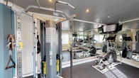 Located at 4874 E Los Coyotes Diagonal in Long Beach, California, this dedicated fitness center is a thoughtfully designed, single-floor facility built to support a comprehensive workout experience. The space features rubber flooring throughout, providing both durability and comfort underfoot during exercise.

The cardio zone is equipped with multiple treadmills and an elliptical trainer, each positioned to take advantage of natural light streaming through large windows fitted with white plantation shutters. Wall-mounted flat-screen televisions accompany the cardio equipment, offering entertainment during workouts.

Strength training is well represented with a multi-station cable weight machine offering a wide range of resistance settings, along with a full rack of dumbbells spanning a broad weight range. A flat utility bench is also available for free weight exercises.

Floor-to-ceiling mirrors line the walls, visually expanding the space and allowing users to monitor their form during workouts. Recessed lighting in the grey-painted ceiling provides consistent illumination throughout the room, complementing the natural light. Crown molding adds a refined architectural touch to the interior.

The space includes a dedicated entry door with an adjacent window, as well as hygiene amenities such as a paper towel dispenser. The overall ambiance is clean, functional, and well-maintained, making it an effective fitness resource.