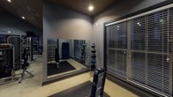 Located at 200 Citygreen Way in Chattanooga, Tennessee, this well-appointed fitness center offers a comprehensive workout environment designed to support a wide range of exercise routines. The space features a dark, dramatic ceiling with recessed lighting and multiple ceiling fans that ensure proper air circulation throughout the facility.

The cardio zone is lined with a row of treadmills, elliptical machines, and stationary bikes, each equipped with individual mounted flat-screen televisions for entertainment during workouts. Large windows along the cardio wall allow natural light to filter in and provide views of the surrounding exterior.

The strength training area is outfitted with a variety of cable and resistance machines, weight benches, and functional training equipment. A dedicated free-weight section includes a tiered medicine ball rack and a full-length wall mirror, ideal for form monitoring during exercises. Rubber flooring mats are strategically placed throughout the workout zones to provide safety and durability.

The overall ambiance is sleek and modern, with cool gray walls, dark accents, and focused lighting creating a motivating atmosphere. A small utility area with cabinetry is also visible within the space. Posted fitness center rules ensure a well-maintained and respectful environment for all users.