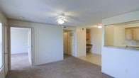 This single-floor residence offers a functional and straightforward layout designed for comfortable everyday living. The main living area is finished with wall-to-wall carpeting that adds warmth underfoot, while a ceiling fan with an integrated light fixture provides both airflow and ambient lighting. The textured ceiling adds a classic architectural touch throughout the space.

The living area flows openly toward the kitchen, which is separated by a half-wall or breakfast bar, creating a semi-open connection between the two spaces. The kitchen features wood-toned cabinetry, countertop workspace, and tile flooring, offering a practical cooking and meal preparation environment that contrasts neatly with the carpeted living zone.

A hallway visible from the living area leads to additional rooms, including what appears to be a closet with louvered or paneled doors, suggesting ample storage options. The neutral color palette of whites and beiges throughout creates a clean, cohesive look that is easy to personalize. Recessed vents are integrated into the ceiling for climate control. Overall, the space presents a well-organized, single-level layout with a relaxed and utilitarian ambiance suited for everyday residential use.