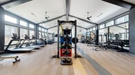 Located on Meadowbrook Pkwy in Colorado Springs, CO, this single-story fitness center is designed to accommodate a full range of workout disciplines within a thoughtfully organized, open layout. The facility is defined by its striking vaulted ceiling supported by bold dark-painted beams, creating an expansive, airy atmosphere enhanced by recessed lighting and ceiling fans throughout.

The main training floor is divided into distinct functional zones. The cardio area lines one side of the space with a row of treadmills and elliptical machines, while the central zone features a multi-use functional training rig equipped with resistance bands, kettlebells, medicine balls, and plyo boxes for dynamic training. A dedicated strength area houses selectorized cable machines, benches, and a comprehensive dumbbell rack, all arranged with ample spacing.

A separate, naturally lit room serves as a studio space for cycling, rowing, and mat-based workouts, featuring large dark-framed windows that flood the area with natural light and views of the surrounding neighborhood. Full-length mirrors are positioned strategically throughout the facility to maximize sightlines and support proper form. The patterned carpet tile flooring adds visual interest while providing comfort underfoot across the entire space.