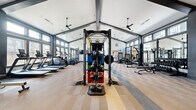 Located on Meadowbrook Pkwy in Colorado Springs, CO, this single-story fitness center is designed to accommodate a full range of workout disciplines within a thoughtfully organized, open layout. The facility is defined by its striking vaulted ceiling supported by bold dark-painted beams, creating an expansive, airy atmosphere enhanced by recessed lighting and ceiling fans throughout.

The main training floor is divided into distinct functional zones. The cardio area lines one side of the space with a row of treadmills and elliptical machines, while the central zone features a multi-use functional training rig equipped with resistance bands, kettlebells, medicine balls, and plyo boxes for dynamic training. A dedicated strength area houses selectorized cable machines, benches, and a comprehensive dumbbell rack, all arranged with ample spacing.

A separate, naturally lit room serves as a studio space for cycling, rowing, and mat-based workouts, featuring large dark-framed windows that flood the area with natural light and views of the surrounding neighborhood. Full-length mirrors are positioned strategically throughout the facility to maximize sightlines and support proper form. The patterned carpet tile flooring adds visual interest while providing comfort underfoot across the entire space.