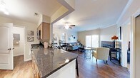 This single-story space showcases an open-concept layout that seamlessly connects the kitchen, dining, and living areas into one cohesive environment. The kitchen is appointed with dark granite countertops that provide a striking contrast against lighter cabinetry, and includes stainless steel appliances. A raised breakfast bar serves as a natural dividing element between the kitchen and the adjoining dining and living spaces.

The living area is warmed by a fireplace framed in stone or tile, adding a focal point to the room. French doors along the far wall invite abundant natural light while offering views to the outdoors. A ceiling fan centered overhead provides both functionality and a finishing architectural touch.

Rich hardwood flooring runs throughout, lending warmth and continuity across the entire open floor plan. Neutral wall tones provide a versatile backdrop throughout the space. The overall layout promotes a comfortable flow between functional areas, making the space well-suited for both everyday living and entertaining.