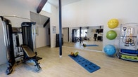 This dedicated fitness studio is designed to support a full range of training activities in a bright, open environment. The space is defined by its striking vaulted ceilings with exposed dark beams, which lend an airy, expansive feel and allow natural light to pour in through high-set windows. The light hardwood flooring adds warmth to the otherwise crisp, white-walled interior, creating an energizing yet welcoming atmosphere.

A large floor-to-ceiling wall mirror spans a significant portion of one wall, making the space feel even more open while providing essential visual feedback during workouts. A structural support column, finished in deep navy, serves as a subtle architectural accent within the room.

The studio is thoughtfully outfitted with a diverse selection of equipment. A multi-station cable and weight machine anchors one side of the room, accompanied by a dedicated weight bench. A neatly organized dumbbell rack offers a full range of free weights for strength training. Cardio and flexibility needs are met with a rowing machine, exercise mat, foam roller, medicine balls, and stability balls. A mounted flat-screen television is also present, ideal for guided workout sessions or entertainment. The space is well-suited for personal training, group fitness, or individual daily exercise routines.