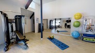 This dedicated fitness studio is designed to support a full range of training activities in a bright, open environment. The space is defined by its striking vaulted ceilings with exposed dark beams, which lend an airy, expansive feel and allow natural light to pour in through high-set windows. The light hardwood flooring adds warmth to the otherwise crisp, white-walled interior, creating an energizing yet welcoming atmosphere.

A large floor-to-ceiling wall mirror spans a significant portion of one wall, making the space feel even more open while providing essential visual feedback during workouts. A structural support column, finished in deep navy, serves as a subtle architectural accent within the room.

The studio is thoughtfully outfitted with a diverse selection of equipment. A multi-station cable and weight machine anchors one side of the room, accompanied by a dedicated weight bench. A neatly organized dumbbell rack offers a full range of free weights for strength training. Cardio and flexibility needs are met with a rowing machine, exercise mat, foam roller, medicine balls, and stability balls. A mounted flat-screen television is also present, ideal for guided workout sessions or entertainment. The space is well-suited for personal training, group fitness, or individual daily exercise routines.