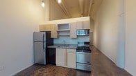 Located at 60 E Spring St in the heart of Columbus, Ohio, this single-floor urban loft showcases a distinctive industrial aesthetic with modern updates. The space is defined by its dramatically high ceilings with exposed structural beams, polished concrete floors, and clean white walls that create an open, airy atmosphere throughout.

The kitchen is efficiently designed and features light wood cabinetry, granite countertops, and a full suite of stainless steel appliances including a gas range, microwave, and refrigerator. A black dishwasher and a single-basin sink complete the functional layout, all set beneath the soaring ceiling that gives the kitchen an expansive feel.

The main living area is generously proportioned and bathed in natural light from large, multi-pane industrial-style windows that frame views of the surrounding downtown Columbus cityscape. The polished concrete flooring continues seamlessly throughout, reinforcing the loft's urban character.

The bathroom is clean and functional, featuring a full tub and shower combination with chrome fixtures and a granite-topped vanity with an undermount sink. A wall mirror and industrial-style lighting complete the space.

The overall ambiance of this unit is a compelling blend of raw, warehouse-inspired architecture and contemporary livability, making it a distinctive offering in the heart of downtown Columbus.