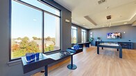 Located at 3500 Cox Rd in Henrico, Virginia, this contemporary amenity lounge offers a well-appointed, open-concept recreation and social space. The room is defined by its modern aesthetic, featuring cool-toned slate-blue walls, light hardwood flooring, and a coffered ceiling with recessed and pendant lighting that adds a refined architectural touch.

Floor-to-ceiling windows line the perimeter, flooding the interior with natural light and providing expansive views of the surrounding Henrico landscape. The space is thoughtfully organized into distinct activity zones. A central billiards table serves as the focal point of the room, accompanied by a shuffleboard table positioned along the windowed wall — both ideal for social recreation. A lounge and dining area, defined by a patterned area rug and round table with modern chairs, offers a comfortable gathering spot. An open shelving unit acts as a subtle divider between zones, adding both function and visual interest.

A wall-mounted flat-screen TV anchors one end of the room, while a glass-partitioned private space — likely a conference or quiet room — is accessible via sliding glass doors. Decorative plants and curated wall art contribute to the polished, welcoming atmosphere. This versatile space is well-suited for both casual socializing and organized events.