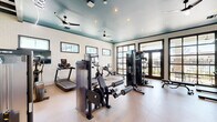 Located at 208 Windfall Way in Myrtle Beach, SC, this fitness center offers a comprehensive workout environment across a single, open floor. The space is defined by its striking teal-painted ceiling accented with exposed wooden beam detailing and neon lighting elements, creating a modern yet inviting atmosphere. White-painted brick walls lend an industrial-chic character throughout, while recessed lighting and ceiling fans ensure the space remains bright and comfortable during workouts.

Natural light flows generously through multiple framed windows positioned along the walls, complemented by large black-framed glass doors that open to an outdoor area, providing views of the surrounding community. The light-toned flooring ties the design together with a clean, polished aesthetic.

The fitness floor is thoughtfully organized to accommodate a wide range of exercise modalities. Cardio equipment includes treadmills, elliptical trainers, a rowing machine, and stationary bikes—several equipped with individual screens. Strength training is well-represented with a multi-station cable machine, weight benches, free weights, and additional resistance equipment. Wall-mounted flat-screen televisions and large mirrors enhance the space further. Built-in shelving and cabinetry along one wall provide practical storage, completing this well-appointed facility.
