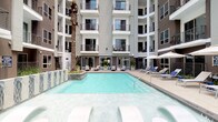 This expansive outdoor amenity space is centered around a resort-style swimming pool and spa, nestled within the open-air courtyard of a contemporary multi-story building. The pool area is framed by clean, light-toned concrete decking and surrounded by a generous selection of lounge chairs and stylish blue-and-white patterned outdoor seating arrangements, creating a relaxed and inviting atmosphere. A tall palm tree anchors the courtyard, adding a tropical accent to the modern architectural backdrop of stacked balconies clad in neutral grey tones with sleek metal railings.

Adjacent to the pool, a covered outdoor kitchen provides an exceptional space for entertaining. Equipped with a stainless steel grill set into a stone-accented counter with a marble-look surface, as well as a utility sink and mounted flat-screen television, this area is designed for both function and leisure. The pergola-style cover overhead offers partial shade while maintaining an open, airy feel with views to the surrounding neighborhood.

Throughout the courtyard, thoughtfully arranged planters with lush greenery soften the hardscape and add vibrancy to the space. The overall design blends upscale finishes with a relaxed outdoor lifestyle, making this amenity area a standout feature of the property.