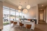 This single-story space is designed with a refined, contemporary aesthetic that balances professionalism with comfort. The reception area greets visitors with light wood-look flooring, a sleek white desk flanked by warm cognac chairs, and a row of four wood-framed art prints displayed along a crisp white wall. Woven globe pendant lights hang overhead, adding texture and warmth to the welcoming atmosphere. Floor-to-ceiling glass windows flood the entry with natural light and offer views of the surrounding outdoor landscape.

The space transitions into a dramatic lounge area characterized by soaring double-height ceilings and an abundance of natural light streaming through tall windows. This area is anchored by a tufted camel leather sofa paired with jewel-toned velvet accent chairs in deep green. A built-in media niche clad in metallic mosaic tile houses a wall-mounted television above slate-blue cabinetry. Living moss wall panels and lush potted dracaena plants bring organic texture throughout, while curated artwork—including a bold abstract canvas—adds dynamic visual interest. The combination of dark tile flooring, layered area rugs, and eclectic decorative accents creates a rich, sophisticated ambiance throughout the entire space.