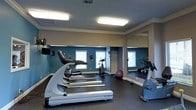 This fitness center offers a dedicated space for exercise and wellness, thoughtfully designed to support a variety of workout routines. The room features durable dark rubber flooring, providing both safety and comfort underfoot during high-intensity training sessions. Crown molding and a textured ceiling add a finished, polished look to the space.

The cardio area is prominently equipped with a row of treadmills, including True-brand models, as well as an upright stationary bike, catering to a range of cardiovascular preferences. A stability ball and additional free-weight equipment round out the available fitness options. Full-length mirrors line the walls, enhancing the sense of space while allowing users to monitor their form during workouts.

Natural light enters through multiple windows fitted with blinds, complemented by bright flush-mounted ceiling fixtures that ensure the room remains well-lit at all times. A wall-mounted television provides entertainment during workouts. The accent walls in a deep teal tone add a modern, energizing aesthetic to the overall environment. The single-floor layout keeps everything easily accessible, making this fitness center a practical and inviting destination for daily exercise.