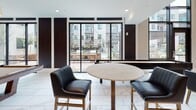 This single-story clubhouse and amenity space is designed with a refined, contemporary aesthetic that blends warm natural materials with modern finishes throughout. The grand entrance lobby immediately impresses with soaring double-height ceilings, floor-to-ceiling black-framed glass walls, exposed wooden ceiling beams, and a striking chandelier overhead. A white grand piano anchors the entry, adding an elegant focal point beside a broad staircase clad in travertine-style stone.

The main lounge area features large-format marble-look porcelain tile flooring, which flows seamlessly through the entire space. Comfortable seating arrangements are set against a dramatic herringbone-patterned feature wall with an integrated flat-screen television, creating an inviting social hub. Floor-to-ceiling windows and sliding glass doors offer expansive views of an outdoor courtyard and resort-style pool.

A fully equipped catering kitchen is outfitted with stainless steel appliances including double wall ovens, granite countertops, rich dark cabinetry, light wood-paneled walls, and deep-toned brick accent tile. Adjacent to the kitchen, a long bar counter with warm gold cabinetry overhead and cream upholstered stools provides additional gathering space.

The game room offers a full-size billiard table, a mounted flat-screen display, a brick-accented bar ledge with counter seating, and direct access to the outdoor amenity deck, completing this well-appointed communal facility.