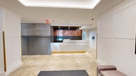 This elegantly designed lobby and hallway space serves as the welcoming centerpiece of a well-appointed residential building managed by First Realty Management. The entrance greets visitors through a set of sleek glass revolving doors framed in black, opening into a generously sized foyer bathed in natural light from expansive floor-to-ceiling windows that offer views of lush greenery outside.

The flooring throughout is a standout feature — light-toned parquet tiles laid in a striking herringbone chevron pattern lend warmth and visual interest to the entire space. A prominent reception desk anchors the lobby, finished in white marble with gold-accented base panels and backed by a rich copper-toned wall feature, creating a refined and contemporary focal point.

Architectural details include white structural columns with gold trim, recessed cove lighting along coffered ceiling trays, and grey wainscoting panels that add texture and depth to the walls. A seating area near the entrance features modern upholstered benches, a round pendant light fixture in gold, and tasteful greenery, fostering a welcoming ambiance.

Additional amenities visible include a resident mailbox bank, a digital information display screen, and a well-lit extended corridor connecting to elevators and further building facilities, completing a lobby that balances functionality with upscale, modern design.