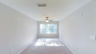This single-floor space offers a clean, functional layout designed with comfort and practicality in mind. The interior is characterized by neutral tones throughout, including soft white walls and light-colored carpeting that lend a calm, airy atmosphere to the space. Large windows fitted with vertical blinds allow generous amounts of natural light to pour in, creating a bright and welcoming environment while framing views of the surrounding greenery outside. A ceiling fan with an integrated light fixture is centrally installed, ensuring efficient air circulation and supplemental lighting. The smooth white ceilings are complemented by clean white baseboards that neatly frame the carpeted floors. HVAC vents are thoughtfully integrated into the ceiling, reflecting a practical approach to climate control throughout the space. The overall design is straightforward and uncluttered, making it highly adaptable for a variety of uses and furniture arrangements. The combination of natural light, neutral finishes, and well-maintained surfaces contributes to a comfortable and inviting ambiance that is both versatile and easy to personalize.