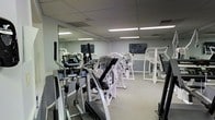 The fitness center at Mission Viejo Villas is a well-appointed, single-floor facility designed to support a comprehensive workout routine. The room is outfitted with a variety of modern exercise machines, including ellipticals equipped with digital displays, a leg press machine, and additional strength training equipment arranged throughout the space. A wall-mounted flat-screen television provides entertainment during workouts, while a full-length mirror along one wall visually expands the room and allows users to monitor their form. The flooring features a neutral carpet suitable for gym use, and the ceiling is finished with a standard drop-tile panel system housing recessed fluorescent lighting that evenly illuminates the entire space. The light gray walls contribute to a clean, functional aesthetic. The layout is open and organized, allowing comfortable movement between machines. Overall, the fitness center presents a practical and functional environment well-suited for residents seeking convenient access to exercise amenities within the Mission Viejo Villas community.