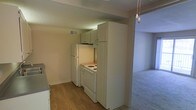 This single-story space features an open layout connecting the kitchen and living area. The kitchen is equipped with white appliances, including a refrigerator and an electric coil range, complemented by light sage-green cabinetry that extends to the ceiling for added storage. Upper cabinets are positioned above the cooking area, maximizing vertical space. The kitchen flooring consists of wood-look hardwood or laminate planks, providing a practical and clean surface. A textured ceiling runs throughout the unit, and a visible HVAC vent ensures climate control. The kitchen transitions seamlessly into the adjacent living area, which features wall-to-wall carpet and neutral-toned walls, creating a cohesive and understated aesthetic. A light fixture with a pull chain is visible at the edge of the living space, providing overhead illumination. The overall design is functional and straightforward, with a neutral color palette throughout that offers a blank canvas for personalization.