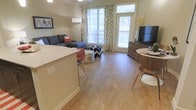 This single-floor unit features an open-concept layout that seamlessly connects the kitchen and living areas, creating a cohesive and functional living environment. Light hardwood-style flooring extends throughout, adding warmth and continuity to the space.

The kitchen includes a breakfast bar with granite-style countertops and bar seating, making it ideal for casual dining or entertaining. The living area is generously proportioned, accommodating a sectional sofa and additional seating with room to spare.

Large windows with decorative leaf-print curtains allow abundant natural light to fill the space, enhancing its airy feel. A glass-paneled door provides access to an outdoor area, offering views of the exterior and further brightening the interior.

The dining area is positioned near a wood-tone media console, creating a natural separation between functional zones. Neutral wall tones complement the flooring and furnishings, contributing to a clean, modern aesthetic. Overall, the space balances practicality with a welcoming ambiance suited to everyday living.
