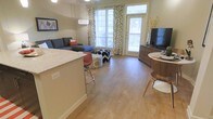 This single-floor unit features an open-concept layout that seamlessly connects the kitchen and living areas, creating a cohesive and functional living environment. Light hardwood-style flooring extends throughout, adding warmth and continuity to the space.

The kitchen includes a breakfast bar with granite-style countertops and bar seating, making it ideal for casual dining or entertaining. The living area is generously proportioned, accommodating a sectional sofa and additional seating with room to spare.

Large windows with decorative leaf-print curtains allow abundant natural light to fill the space, enhancing its airy feel. A glass-paneled door provides access to an outdoor area, offering views of the exterior and further brightening the interior.

The dining area is positioned near a wood-tone media console, creating a natural separation between functional zones. Neutral wall tones complement the flooring and furnishings, contributing to a clean, modern aesthetic. Overall, the space balances practicality with a welcoming ambiance suited to everyday living.