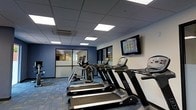 Located at 646 W Bay Street in the heart of Savannah, Georgia, this fitness center offers a well-appointed space designed to support an active lifestyle. The room features a dropped ceiling with recessed panel lighting that provides consistent, even illumination throughout the space. The flooring is covered in a patterned, multi-toned carpet that adds a modern touch while providing comfort underfoot. The walls are painted in a combination of crisp white and bold navy blue accents, giving the space a clean, professional aesthetic. A wall-mounted flat-screen television is positioned for easy viewing during workouts, enhancing the overall exercise experience. The cardio area is equipped with a row of treadmills and elliptical machines, offering a variety of options for cardiovascular training. Large interior windows and glass partitions along one wall allow for sightlines into an adjacent area, contributing to an open, connected feel. A ceiling-mounted HVAC unit ensures climate control and comfort during use. The overall ambiance is functional and modern, making this fitness center a practical and well-equipped amenity for occupants of the building.