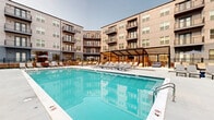 This outdoor amenity space at 4455 Condor Dr in Hilliard, Ohio, is a well-appointed resort-style pool courtyard nestled within a modern multi-story residential complex. The centerpiece is a generously sized swimming pool with crystal-clear blue water, entry handrails, and clearly marked depth indicators, designed to accommodate both leisurely swims and relaxed lounging. The expansive pool deck surrounds the water on all sides and is furnished with an extensive collection of cushioned chaise lounge chairs and umbrellas, offering ample space for sunbathing and outdoor relaxation. A pergola structure anchors one end of the courtyard, providing a shaded retreat and a focal point for the overall layout. String lights and landscape plantings add warmth and visual interest to the space, enhancing its inviting ambiance during evening hours. The courtyard is enclosed by the surrounding multi-story building, featuring modern facades with large windows, private balconies, and clean architectural lines that frame the space on multiple sides. A secure perimeter fence defines the pool area while maintaining an open, airy feel. The ground-level amenity spaces visible through glazed storefronts suggest access to additional interior resident amenities. The entire courtyard benefits from abundant natural light and open sky views above.