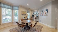 Located at 100 Preston Woods Trail in Sandy Springs, Georgia, this single-floor amenity space is thoughtfully designed to serve as a functional and welcoming community gathering area. The interior is finished with neutral warm-gray walls, crown molding, and light travertine tile flooring that lends a clean, polished aesthetic throughout.

The main area centers around a round wooden pedestal dining table surrounded by sleek wooden chairs, all anchored by a colorful woven area rug that adds a vibrant, eclectic touch to the space. A striking bay window floods this seating area with natural light and offers views of the surrounding greenery, complemented by patterned Roman shades that add a decorative accent.

Along one wall, a built-in counter-height workstation provides a dedicated business center equipped with computers, a printer, and bar-style seating — ideal for residents who need a productive workspace. Decorative wall art, including abstract paintings and a sculptural metallic plate arrangement, adds an artistic dimension to the room.

A corridor with additional colorful artwork leads to an entryway with double interior doors, extending the usable area of the space. Recessed lighting throughout ensures even illumination, while multiple windows on opposing walls maintain a bright, airy ambiance. The overall design strikes a balance between comfort and functionality.