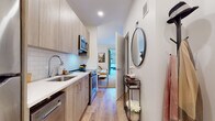 Located at 25 Maple Ave in New Rochelle, NY, this thoughtfully designed single-floor residence offers a well-appointed layout with modern finishes throughout. The galley-style kitchen is a standout feature, outfitted with light wood cabinetry, white quartz countertops, a subway tile backsplash, and a full suite of stainless steel appliances including a refrigerator, range, microwave, and dishwasher. Light hardwood flooring runs continuously through the space, lending warmth and visual cohesion. The entryway transitions smoothly from the kitchen, accented by a circular mirror and a slim console table, creating a welcoming first impression. The bedroom is a calm and comfortable retreat, featuring neutral walls, soft lighting, and space for a full bed, complemented by tasteful décor and a vibrant landscape artwork above the headboard. The bathroom is finished with light marble-style tile flooring and clean white walls, maintaining the home's fresh, minimal aesthetic. A private outdoor patio provides a quiet exterior space with a concrete tile floor, privacy fencing, and room for outdoor furnishings. The overall ambiance of this residence is clean, contemporary, and functional, making it an ideal urban dwelling in the heart of New Rochelle.