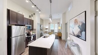 This thoughtfully designed one-bedroom, one-bathroom residence in Austin, TX offers an efficient yet open layout that maximizes every square foot. Soaring ceilings throughout create an airy, expansive feel well beyond its compact footprint.

The kitchen is a standout centerpiece, featuring rich dark cabinetry, stainless steel appliances including a refrigerator, microwave, and range, a tile backsplash, and a generously sized island with a white quartz countertop and undermount sink. A combination of track lighting and modern pendant fixtures illuminate the space beautifully. The kitchen flows seamlessly into the living room, which benefits from natural light streaming through large windows dressed with elegant draping curtains and a ceiling fan overhead.

The bedroom is a serene retreat, showcasing soft blue accent walls, tall windows with outdoor views, and plush carpeted flooring. It connects directly to the full bathroom, which includes a dark wood vanity with a white countertop and an adjacent walk-in closet outfitted with built-in shelving and hanging rods for ample storage.

Wood-look flooring runs throughout the main living areas, tying the open-concept space together with warmth and continuity. This residence combines modern finishes with functional design for comfortable Austin living.
