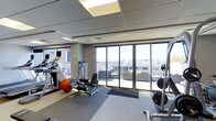 This fitness center is designed to provide a functional and comfortable workout environment. The space is bathed in natural light, thanks to expansive floor-to-ceiling windows that line one wall, offering panoramic views of the surrounding cityscape and creating an open, airy atmosphere. A glass sliding door provides direct access to an outdoor terrace area, further enhancing the connection between the interior workout space and the exterior environment.

The ceiling features a recessed drop-tile design with integrated lighting panels, contributing to even, well-distributed illumination throughout the room. The flooring combines carpet and rubberized mat surfaces, providing appropriate traction and cushioning for various workout activities.

The equipment layout is thoughtfully arranged, including cardio machines such as treadmills and an elliptical trainer positioned near the windows. Strength training equipment, including a cable machine, weight bench, and free weights, are also present. A stability ball adds versatility for functional training. The neutral color palette of gray and beige tones, combined with the abundance of natural light, creates an energizing yet clean and professional workout environment.