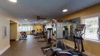 This fitness center is a fully outfitted exercise facility designed to accommodate a wide range of workout routines. The space is organized across a single open floor plan, with distinct zones dedicated to cardiovascular training and strength work. The main area houses a robust selection of Life Fitness equipment, including treadmills, elliptical trainers, stationary and recumbent bikes, and a cable resistance machine, all arranged to allow for comfortable movement between stations. A dedicated free weights section features a tiered dumbbell rack stocked with multiple weight increments, accompanied by a flat weight bench. Full-length mirrors line one wall, enhancing the sense of openness and allowing users to monitor their form. A secondary room provides additional space for flexible exercise, equipped with a recumbent bike, stability ball, and stretching area. The interior features warm yellow-toned walls, recessed lighting, and ceiling fans throughout, contributing to a bright and comfortable environment. Multiple windows with horizontal blinds admit natural light and offer views of the surrounding greenery. Restroom facilities are accessible directly from the fitness floor, and the rubber-floored surfaces throughout provide safe, cushioned footing for all types of exercise.