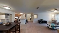 This contemporary apartment showcases an open floor plan that seamlessly integrates the kitchen, dining, and living areas. The kitchen is equipped with wood cabinetry and a convenient breakfast bar, perfect for casual dining or entertaining. Stainless steel appliances complement the space, offering both functionality and style. The living area is expansive, with plush carpeting that extends throughout, creating a cozy atmosphere. Recessed lighting and a central ceiling fixture provide ample illumination. A glimpse of the entryway reveals additional storage options. The neutral color palette, predominantly in shades of white and beige, creates a bright and airy ambiance, making the space feel larger and more inviting. Large openings between areas maintain a sense of continuity and flow, ideal for modern living and socializing.